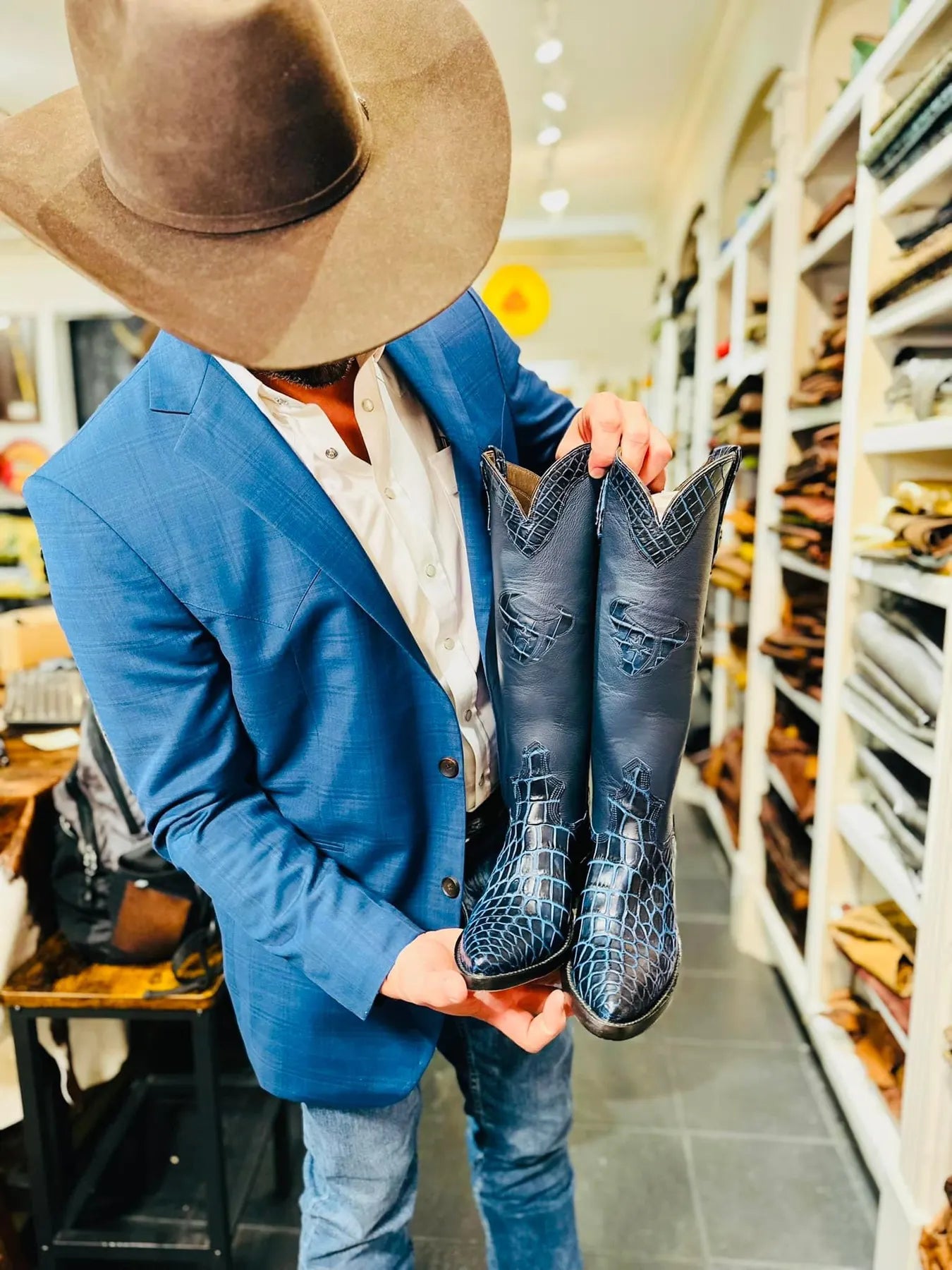 Man in a blue blazer and cowboy hat holding a pair of blue custom boots